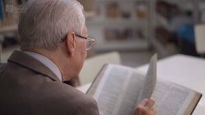 Homem idoso lendo um livro na biblioteca, focado na página. Fundo desfocado com estantes de livros.