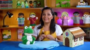 Mulher sorrindo com fantoche de sapo e casa em miniatura em mesa, cenário colorido ao fundo com bonecos.