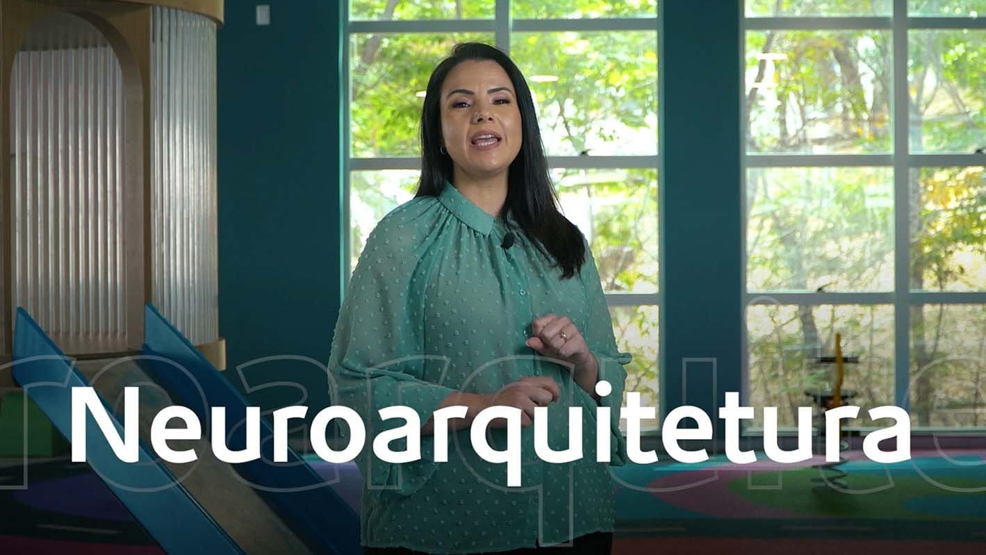 Mulher falando sobre neuroarquitetura em espaço interno iluminado com janelas grandes ao fundo.