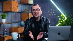 Homem em ambiente de escritório, sentado diante de um laptop com caneca de café, sorrindo para a câmera.