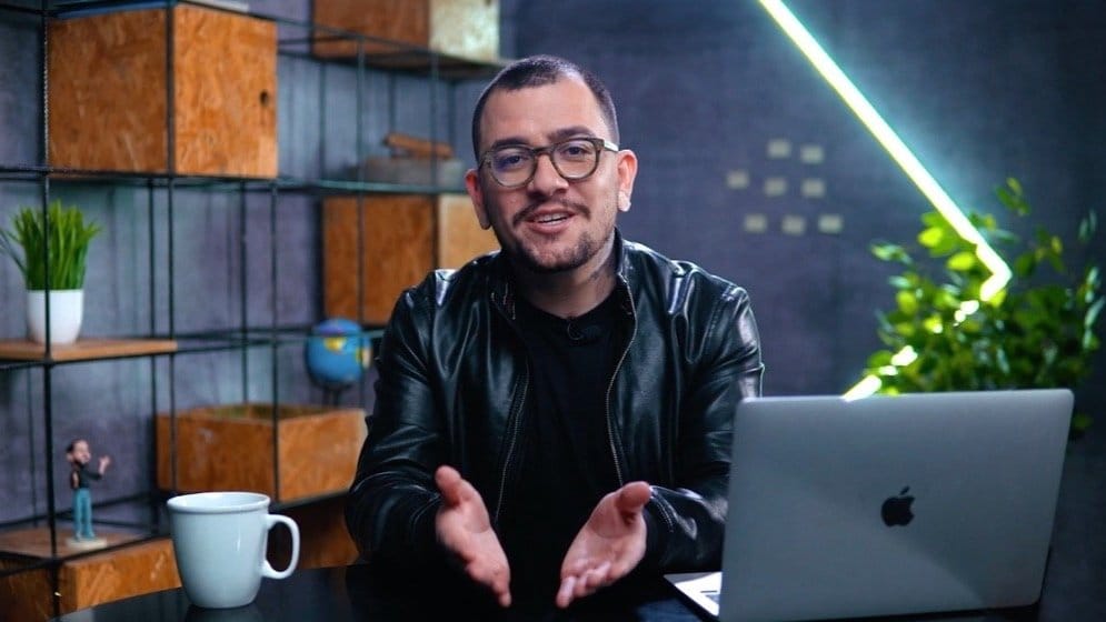 Homem em ambiente de escritório, sentado diante de um laptop com caneca de café, sorrindo para a câmera.