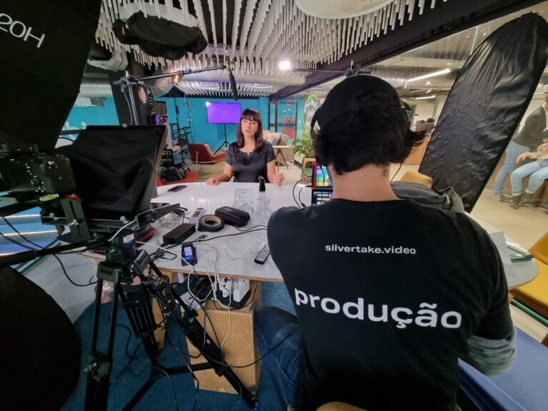 Gravação de vídeo em estúdio com equipe de produção e pessoa apresentando em mesa com equipamentos.