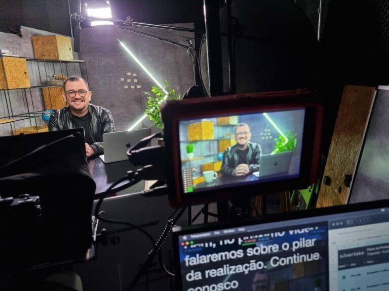 Estúdio de gravação com homem sorrindo, sentado em frente a um laptop, visto através de uma câmera profissional.