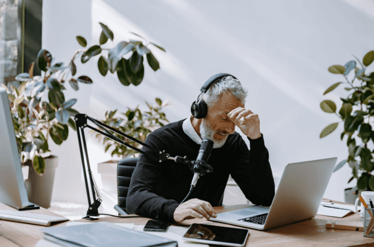 Homem de fones e microfone, com mão na testa, olhando para laptop em escritório com plantas.