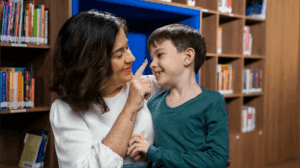 Mulher e criança sorrindo em biblioteca, cercados por prateleiras de livros, compartilhando um momento feliz e educativo.