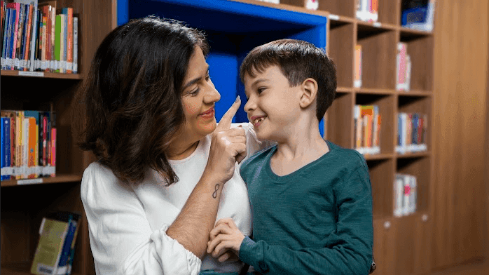 Mulher e criança sorrindo em biblioteca, cercados por prateleiras de livros, compartilhando um momento feliz e educativo.