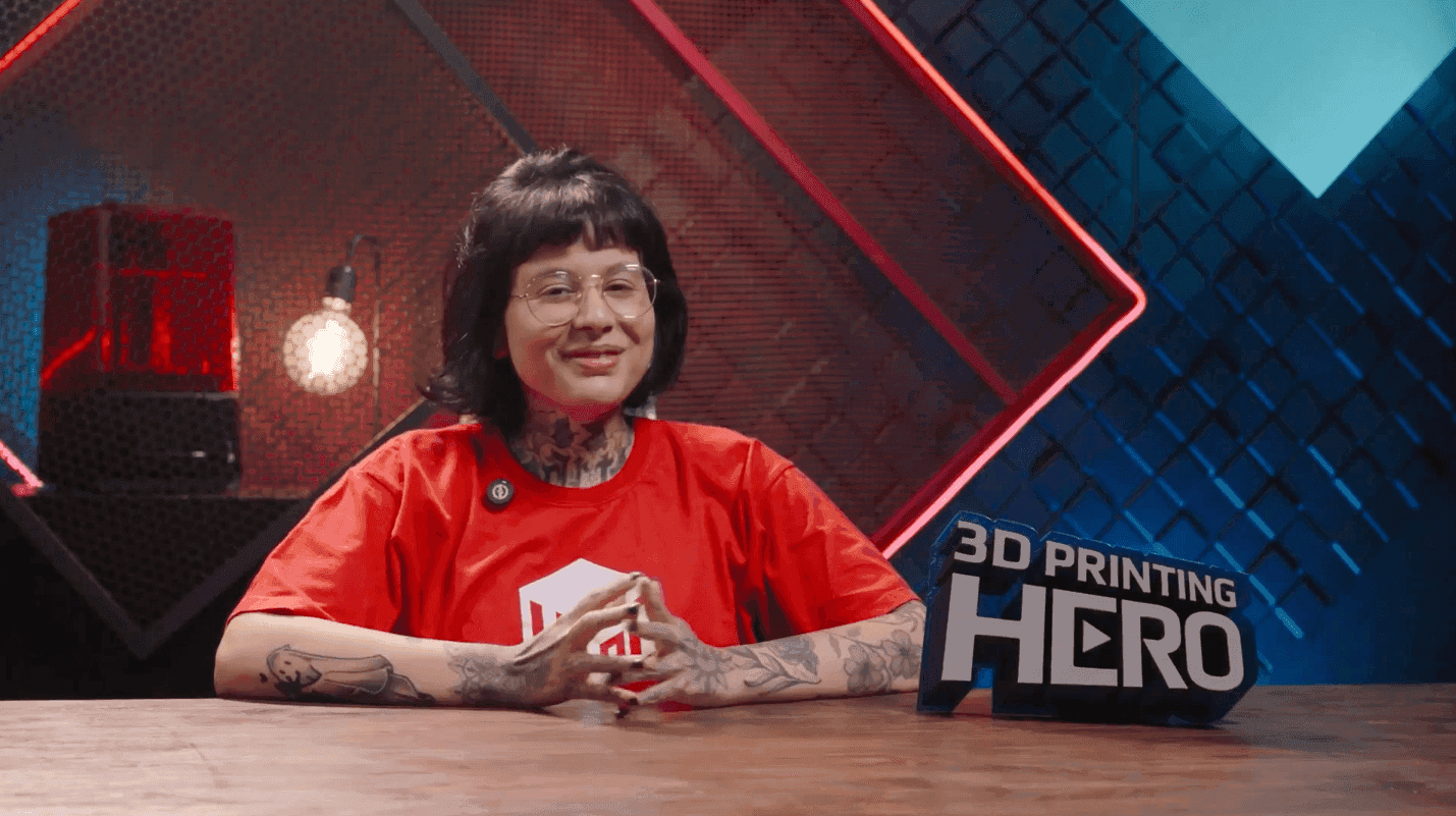 Pessoa sorridente com tatuagens e camiseta vermelha em estúdio de tecnologia com sinal 3D PRINTING HERO.