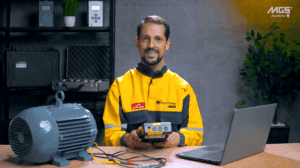 Homem com uniforme amarelo usando dispositivo de medição ao lado de motor elétrico em bancada de oficina.