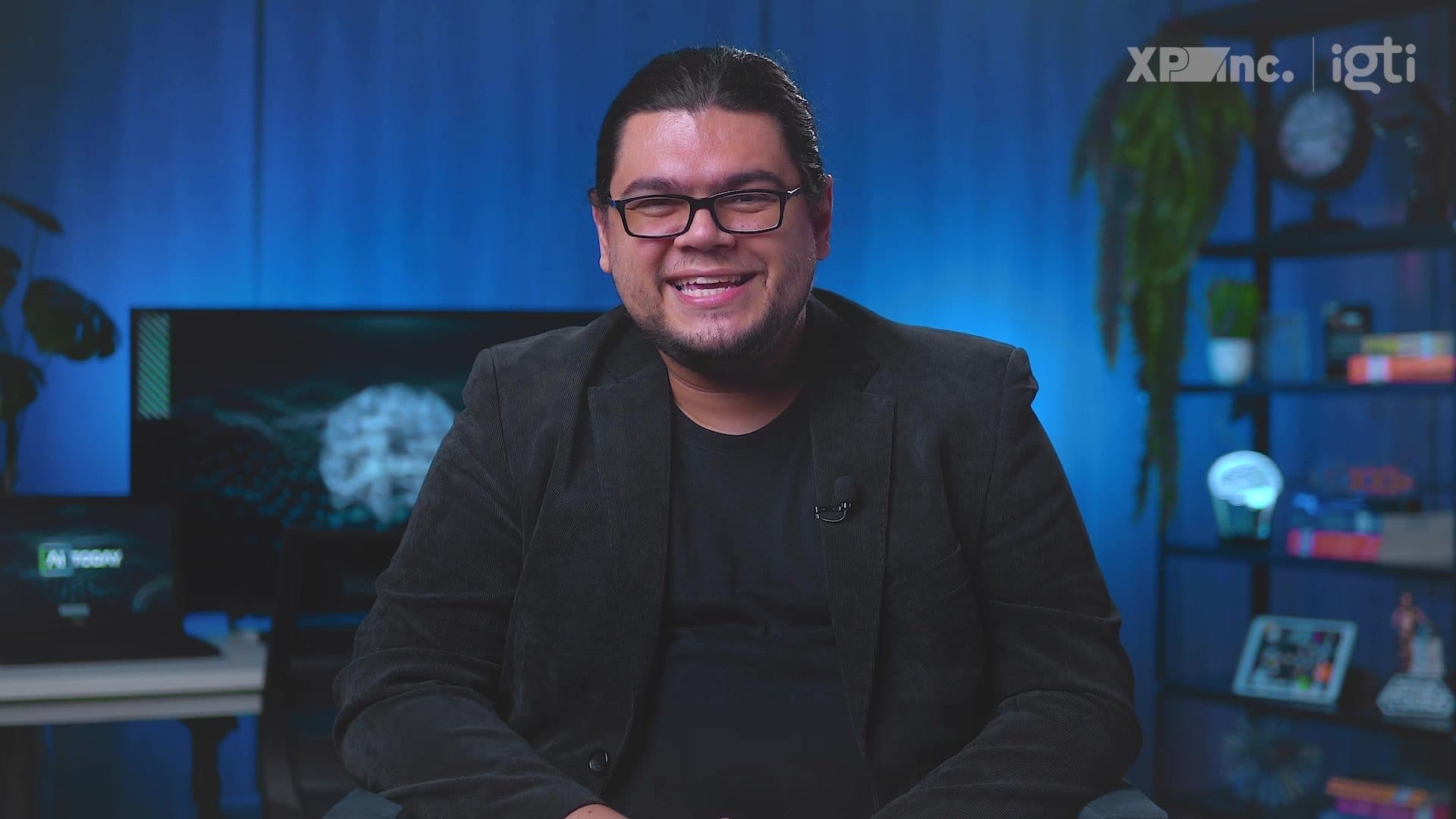 Homem sorrindo em um estúdio com fundo azul, computadores e plantas. Escritório moderno e ambiente iluminado.