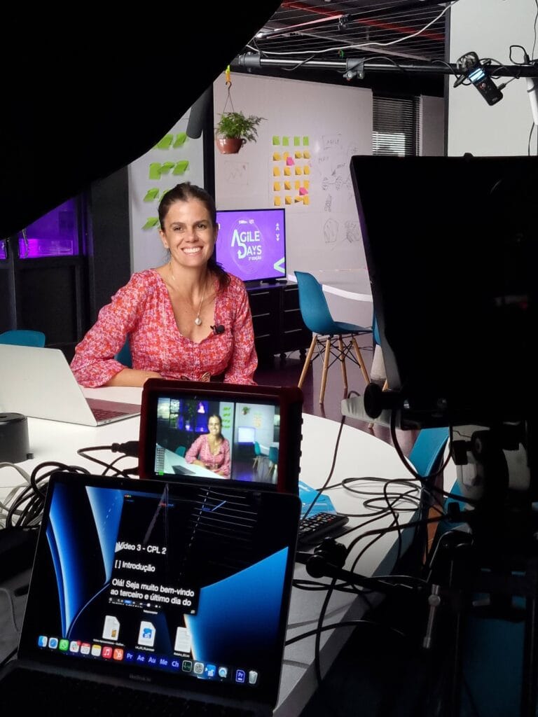 Mulher sorrindo em estúdio de gravação com equipamentos e tela ao fundo. Post-its decoram a parede.