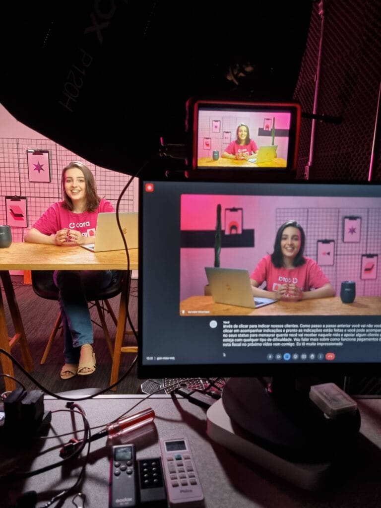 Mulher apresentando vídeo no estúdio com laptop e câmera, imagem sendo exibida em dois monitores, fundo rosa.