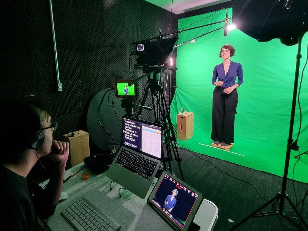 Mulher em estúdio de filmagem com fundo verde, ajustando gravação em câmera profissional e assistente observando.