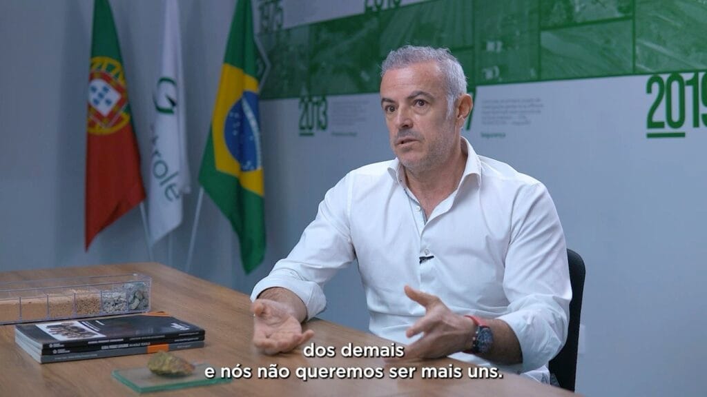 Homem em vídeo institucional em escritório com bandeiras de Portugal e Brasil ao fundo. Ele gesticula enquanto fala.