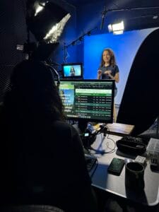 Mulher grava vídeo em estúdio profissional com iluminação, câmera e tela de apresentação ao fundo.