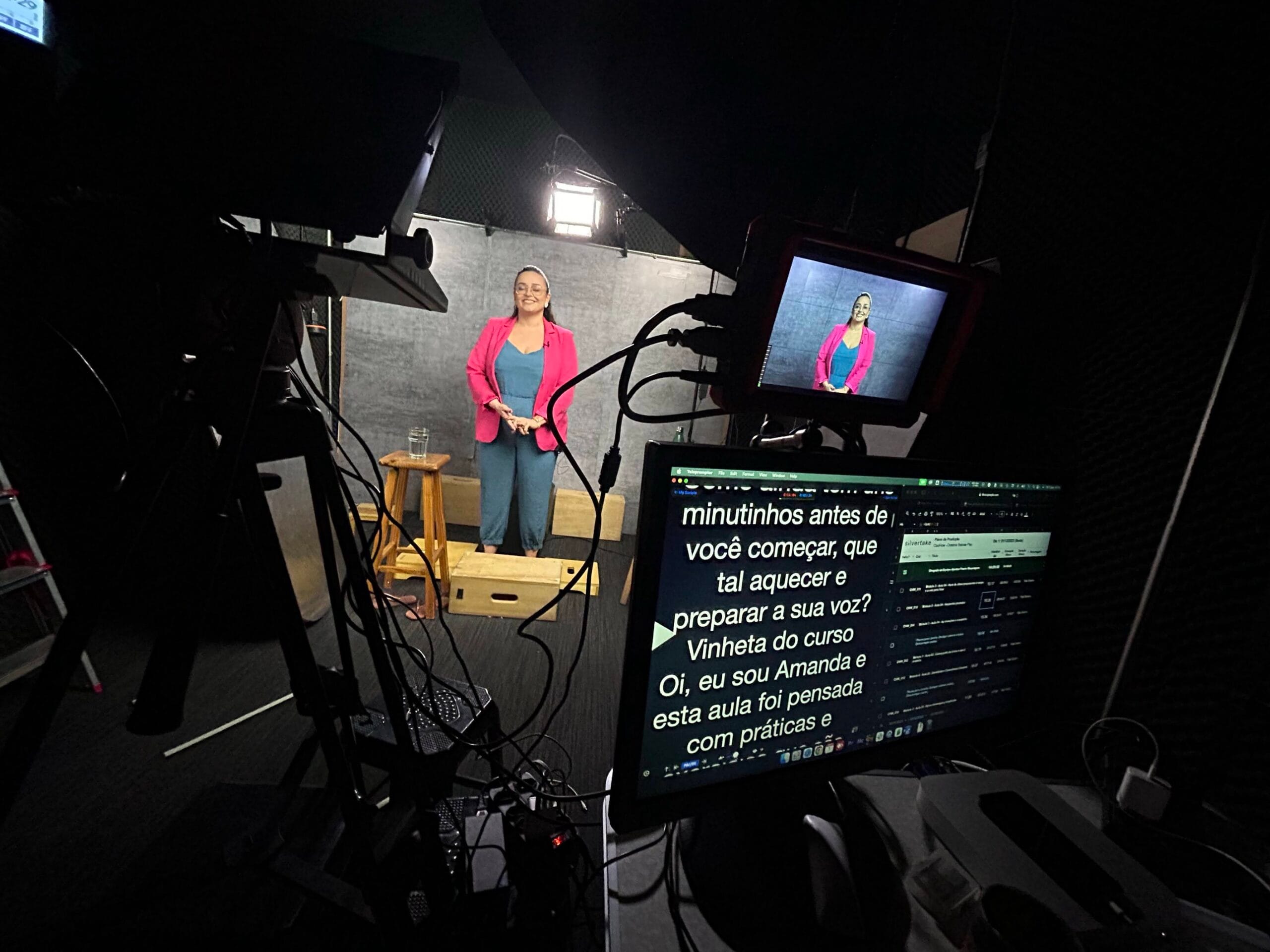 Mulher apresentando em estúdio com teleprompter e equipamentos de filmagem visíveis ao fundo.