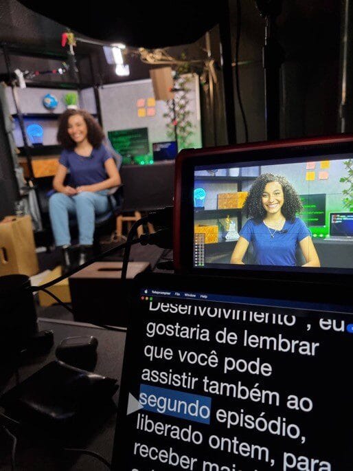 Mulher sorrindo em estúdio de gravação com teleprompter e monitor de vídeo em exibição ao fundo.