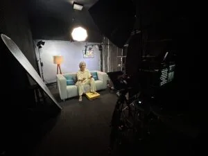 Usuário sentado em um estúdio com iluminação profissional, câmeras e painel acústico, preparado para gravação.
