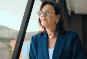 Mulher de óculos e blazer azul olha pensativa pela janela.