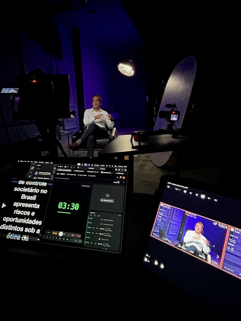 Bastidores de gravação em estúdio com homem sendo filmado e telas em primeiro plano mostrando teleprompter e monitor da câmera