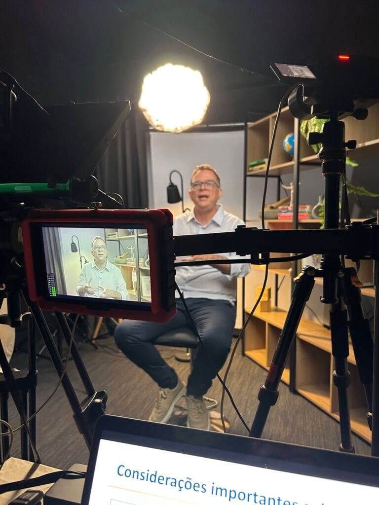 Bastidores de gravação em estúdio, com câmeras, iluminação e monitor exibindo um homem de óculos palestrando