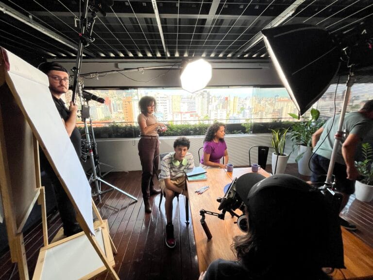 Grupo de pessoas em estúdio preparando gravação, com vista de cidade ao fundo e equipamentos de filmagem.