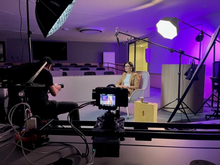 Gravação de vídeo em estúdio com apresentadora, câmera e equipamentos de iluminação. Ambiente moderno e profissional.