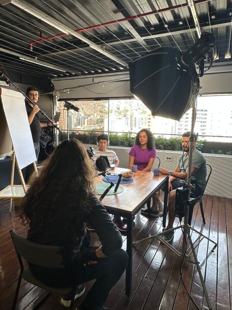 Reunião de produção audiovisual em espaço moderno, com quatro pessoas discutindo e equipamentos de filmagem ao redor.