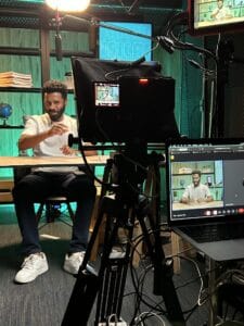Homem sentado em estúdio, gravando conteúdo, com câmeras e equipamentos de áudio ao fundo. Ambiente moderno e funcional.