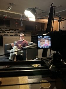 Entrevista em estúdio com homem de óculos sentado no sofá, com iluminação profissional e câmera em foco.