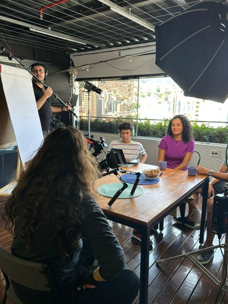 Gravação de vídeo com equipe técnica e participantes em ambiente interno, com vista da cidade ao fundo.