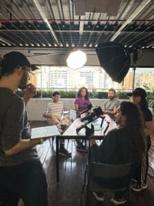 Grupo discutindo em estúdio com equipamentos de filmagem, luzes e laptop em ambiente urbano iluminado.