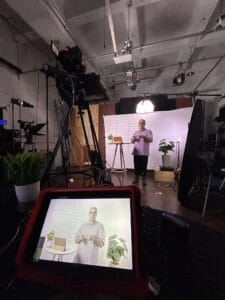 Apresentador em estúdio gravando vídeo, com plantas e equipamentos de iluminação ao fundo, capturado por uma tela.