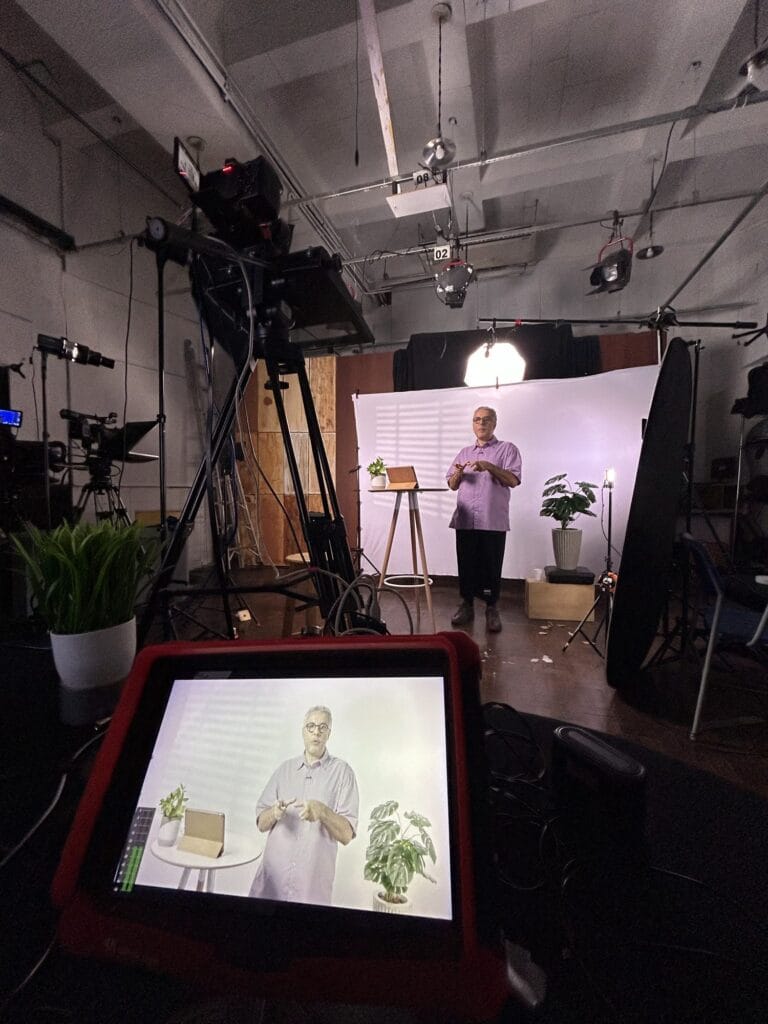 Apresentador em estúdio gravando vídeo, com plantas e equipamentos de iluminação ao fundo, capturado por uma tela.