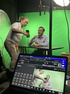 Cenário de gravação com fundo verde, apresentador e assistente usando um celular e laptop para produção de vídeo.