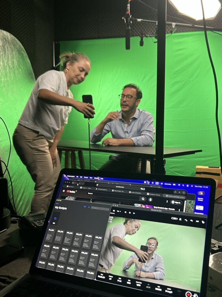 Cenário de gravação com fundo verde, apresentador e assistente usando um celular e laptop para produção de vídeo.