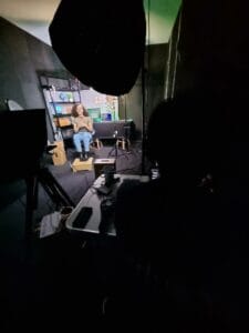 Mulher sentada em estúdio, gravando conteúdo. A cena mostra luzes e equipamentos de produção.