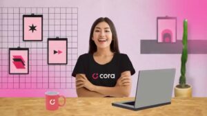 Imagem de mulher sorridente com camiseta da Cora, sentada em uma mesa com laptop e caneca, em ambiente moderno e criativo.