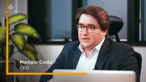Marcelo Costa, CEO da Via Capital, conversando em escritório moderno, próximo a plantas e com laptop à frente.